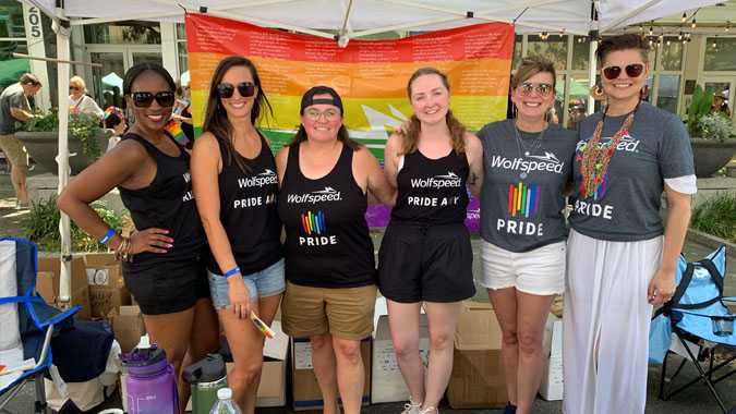 Wolfspeed employees attending the OUT! Raleigh Pride Event standing in front of a pride flag and behind a table with the Wolfspeed logo.
