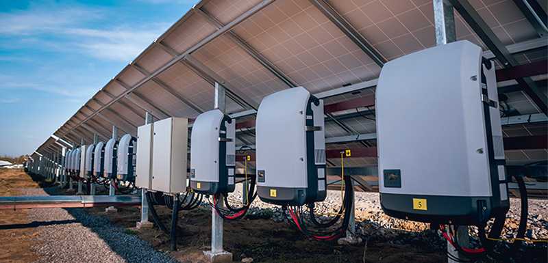 Image of multiple utility-scale string inverters in a line under solar panels.